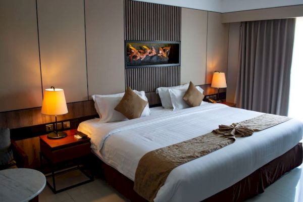 A modern hotel room features a neatly made bed with accent pillows, warm lighting, a wall-mounted fireplace, and a window with closed curtains.
