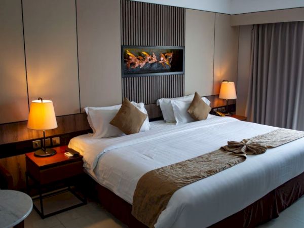 A modern hotel room features a neatly made bed with accent pillows, warm lighting, a wall-mounted fireplace, and a window with closed curtains.