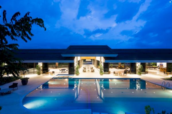 The image shows a luxurious outdoor pool area with a modern building in the background, lit up beautifully under a twilight sky.