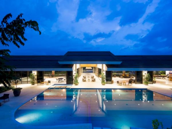 The image shows a luxurious outdoor pool area with a modern building in the background, lit up beautifully under a twilight sky.