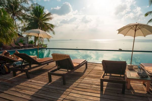A serene seaside pool deck with lounge chairs, umbrellas, and palm trees, offering a picturesque view of the ocean and sky.