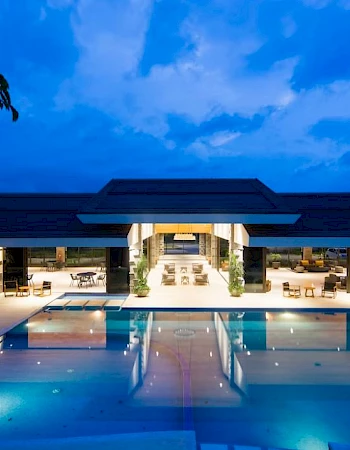 A luxurious, modern building with an illuminated swimming pool at night, surrounded by lounge chairs and trees, under a blue sky.
