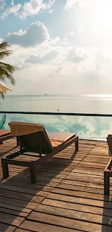 A poolside view with lounge chairs, umbrellas, and palm trees overlooking a serene ocean under a partly cloudy sky and sun.