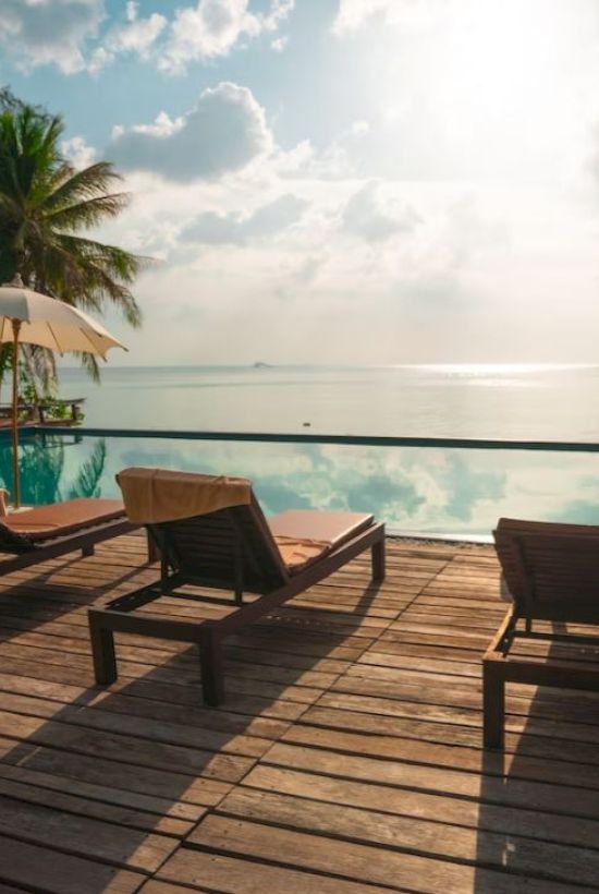 Beachside pool with sun loungers, umbrellas, palm trees, and a serene ocean view under a partially cloudy sky at sunset or sunrise, ending the sentence.