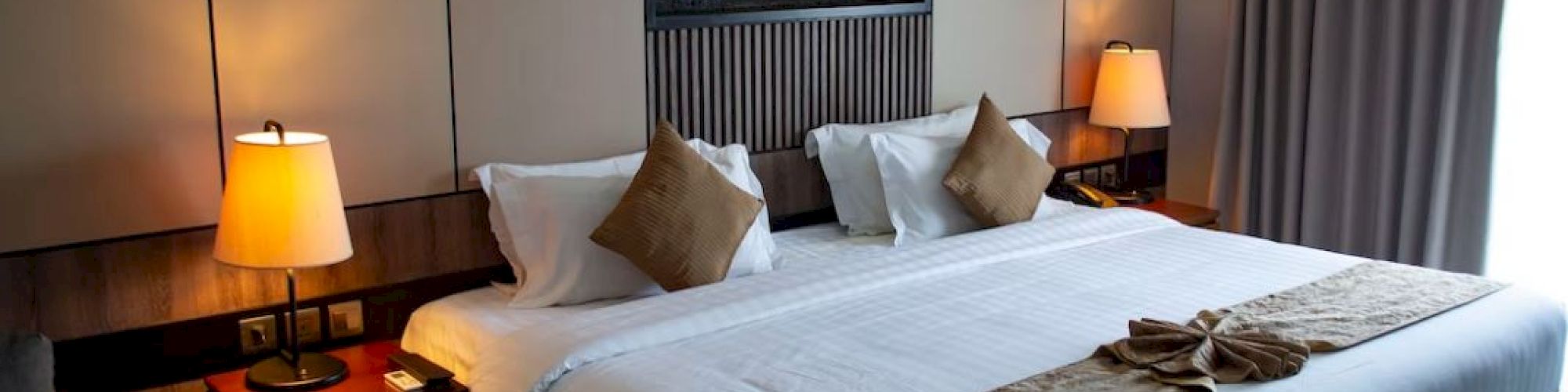 A tidy hotel room features a large bed with pillows, two side tables with lamps, a small table, and a wall-mounted fireplace, ending the sentence.