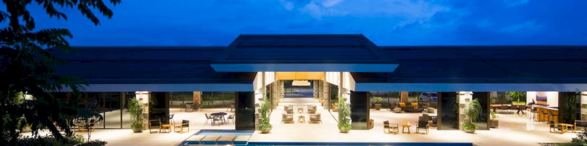 An illuminated outdoor swimming pool with an adjacent building, surrounded by lounge chairs and furniture, under a twilight sky ending the sentence.