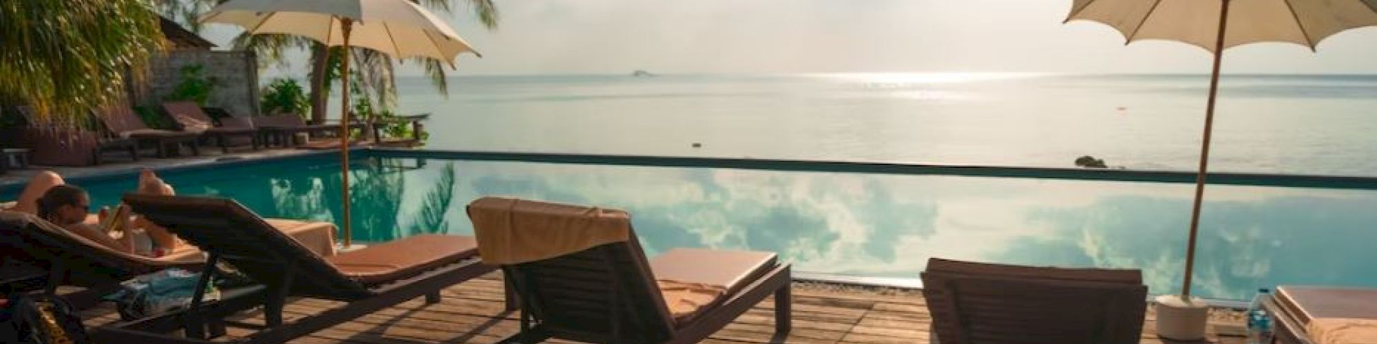 Lounge chairs and umbrellas are placed on a wooden deck overlooking a tranquil infinity pool with a clear view of the ocean and palm trees.