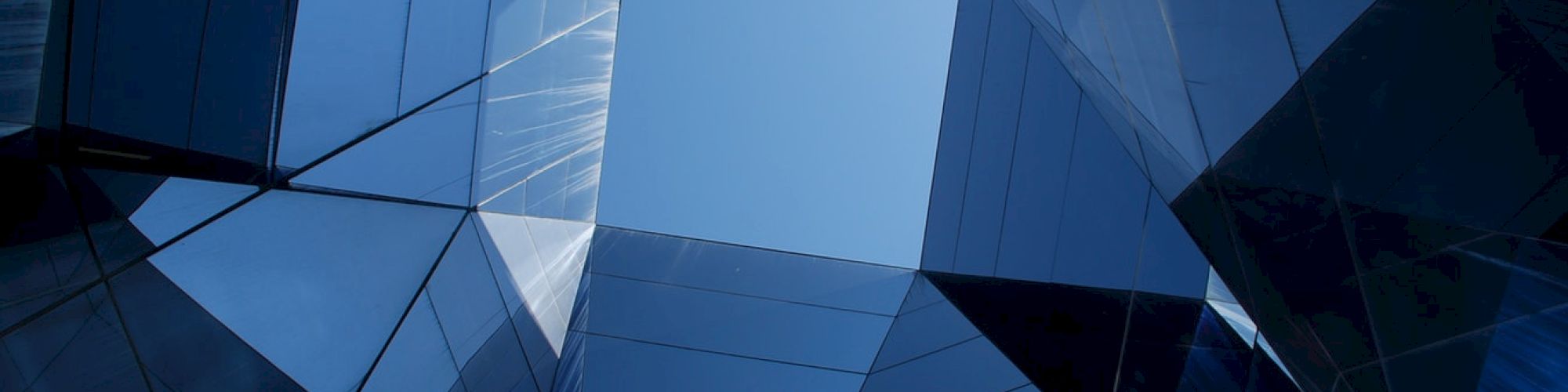 A view looking up through a geometric, glass-covered building structure, revealing a patch of blue sky at the center.