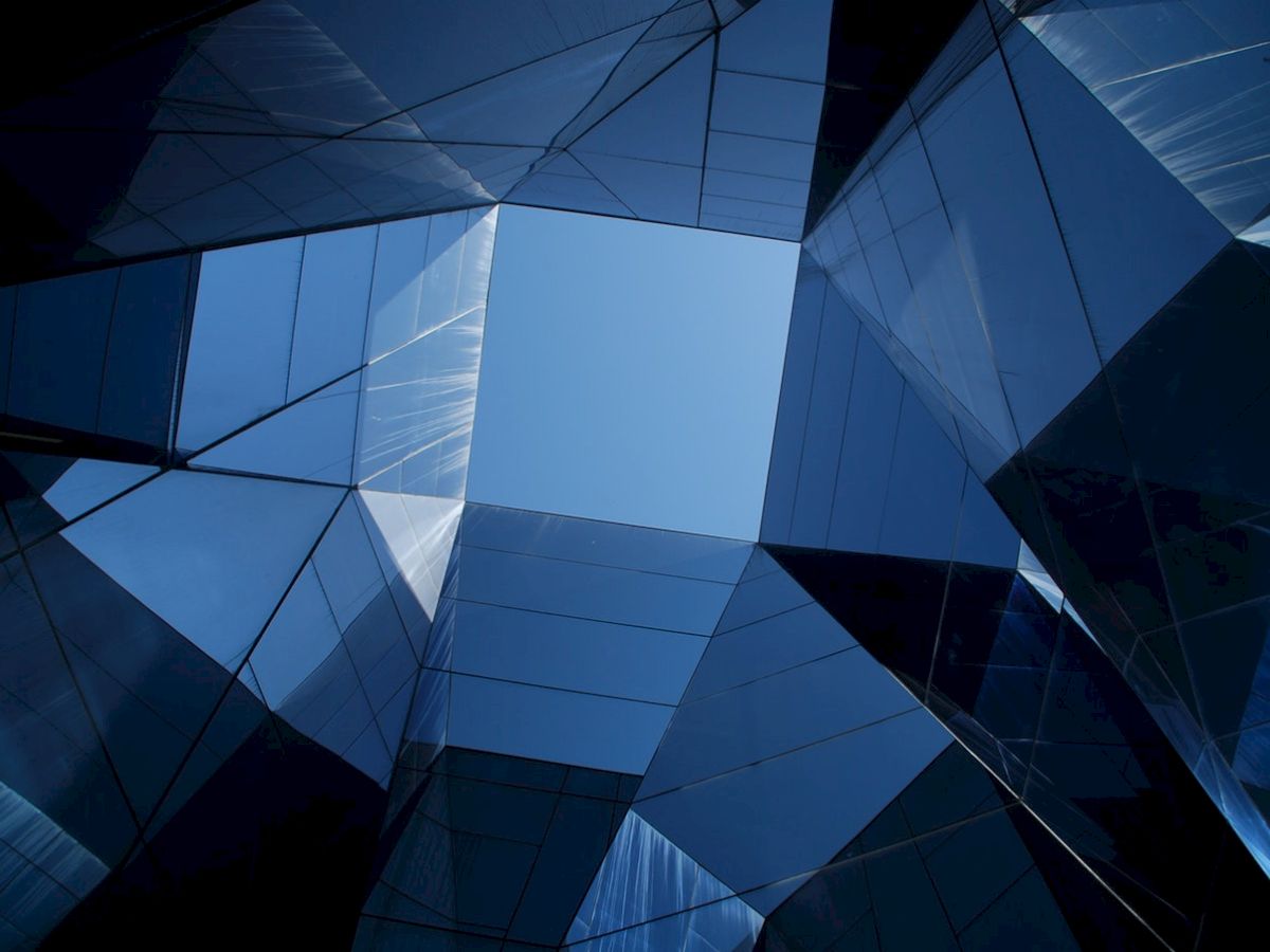This image shows an upward view of a modern glass building with reflective surfaces forming an open geometric pattern, revealing the blue sky.