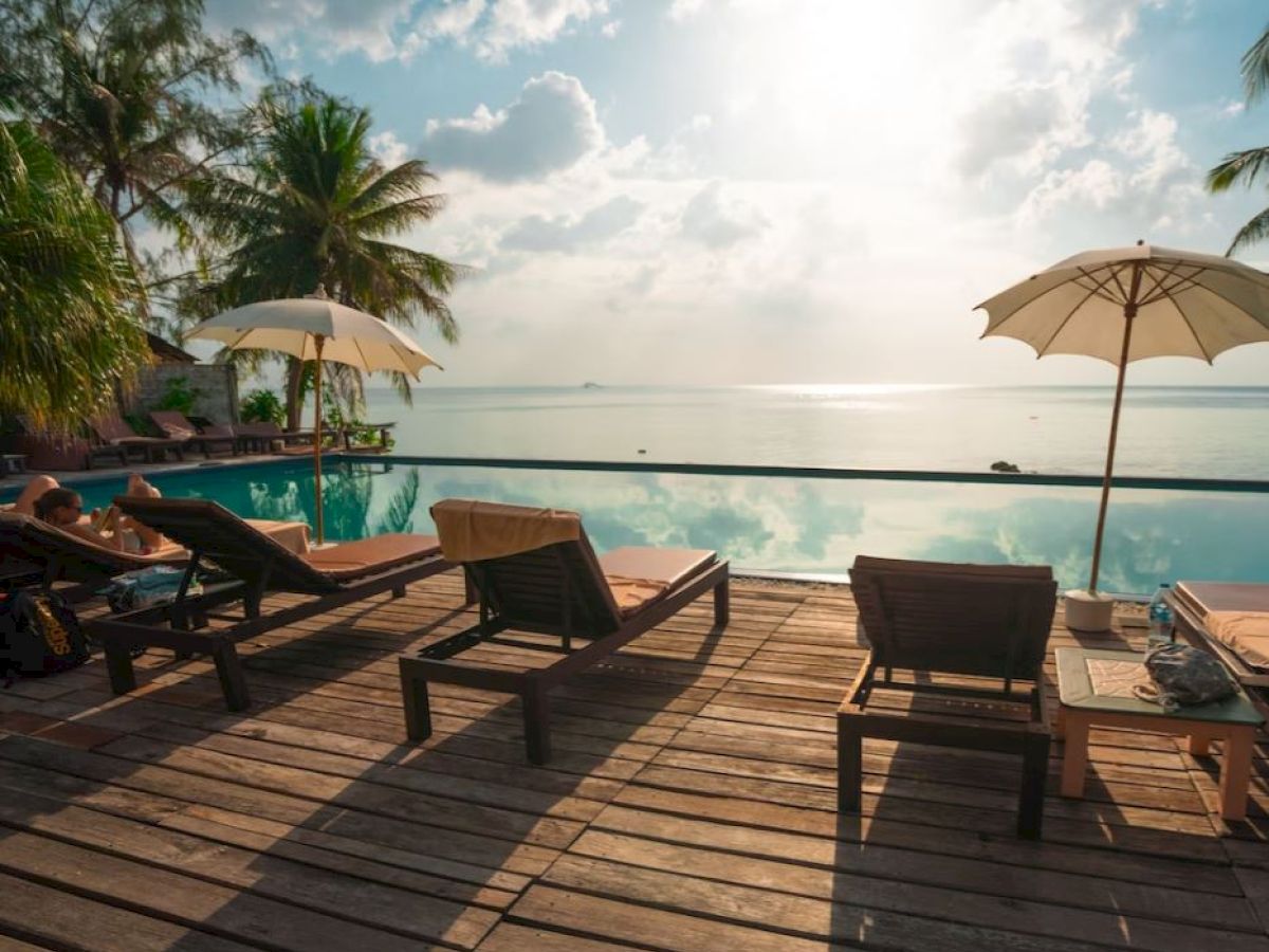 An idyllic poolside scene with lounge chairs, parasols, and a wooden deck overlooking a serene ocean view, under a partly cloudy sky.