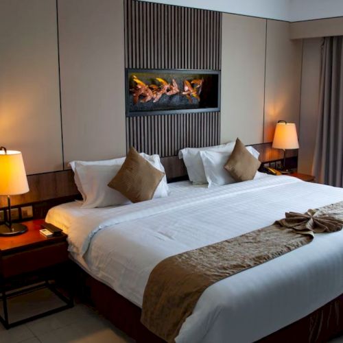 A well-lit hotel room features a large bed with pillows, bedside tables with lamps, a wall-mounted decor piece, and a window with curtains.