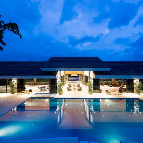 A luxurious building with an expansive outdoor swimming pool, illuminated at dusk, surrounded by seating areas and greenery, under a dramatic sky.