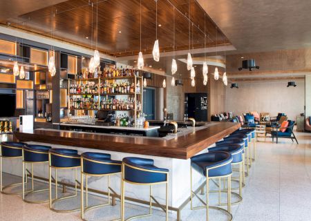 A modern bar with a wooden counter, blue barstools, hanging lights, and shelves stocked with bottles, located in a spacious, well-lit room.
