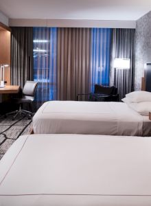 A modern hotel room with two beds, a TV, a desk with a chair, and minimalist decor. The room has large windows with curtains and a stylish design.