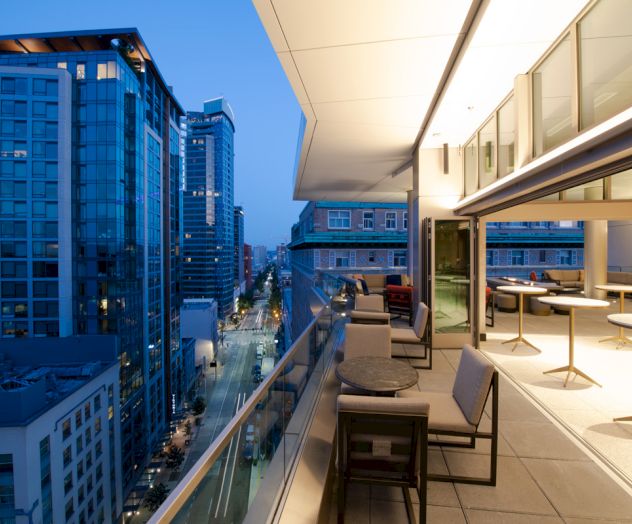 An urban balcony at dusk with modern seating overlooks a cityscape of high-rise buildings and illuminated streets.