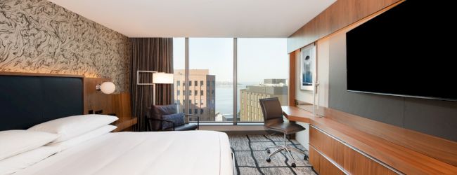 A modern hotel room features a large bed, a wall-mounted TV, a desk with a chair, and a window with a cityscape view in the background.