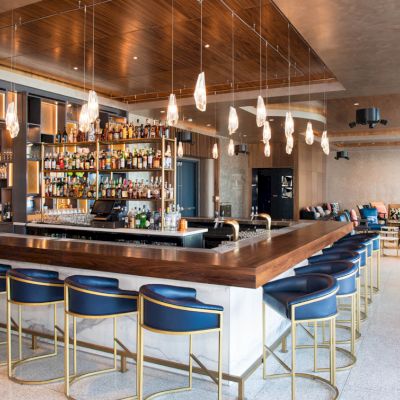 The image shows a modern bar with a counter, high chairs, hanging lights, and shelves stocked with bottles, adjacent to a lounge area with seating.