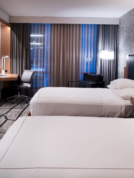 A modern hotel room featuring two neatly made beds, a wall-mounted TV, a desk with a chair, and contemporary decor with patterned wallpaper and floor lamps.