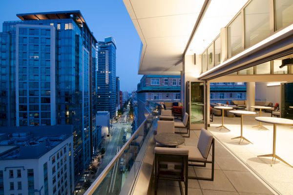 The image shows a modern balcony overlooking a city street lined with high-rise buildings at dusk, with tables and chairs arranged for outdoor seating.