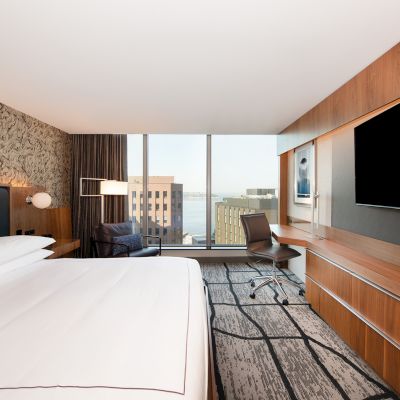 A modern hotel room features a large bed, wall-mounted TV, artwork, desk with chair, a seating area with a view of buildings outside.