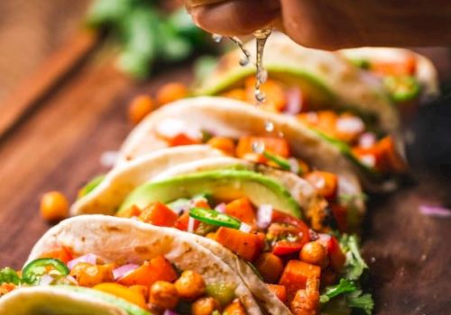 The image shows several tacos filled with vegetables and chickpeas on a wooden board, with a hand squeezing lime juice over them.