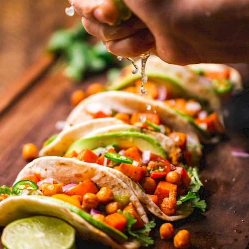 The image shows several tacos filled with vegetables and chickpeas on a wooden board, with a hand squeezing lime juice over them.