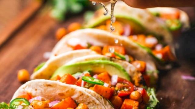 The image shows several tacos filled with vegetables and chickpeas on a wooden board, with a hand squeezing lime juice over them.