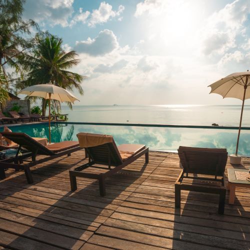 A serene deck with sun loungers and umbrellas overlooks a tranquil infinity pool and a beautiful ocean view under a bright sun and scattered clouds.