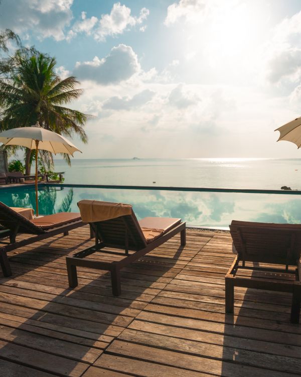 A serene deck with sun loungers and umbrellas overlooks a tranquil infinity pool and a beautiful ocean view under a bright sun and scattered clouds.