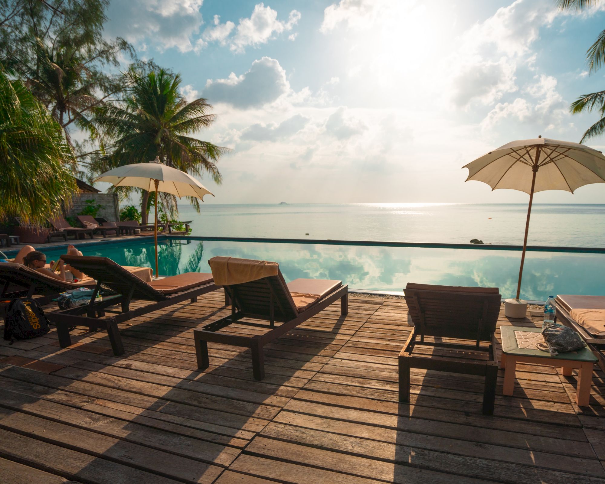 A serene beachside scene with lounge chairs, umbrellas, and palm trees overlooking a calm ocean with a bright sky.