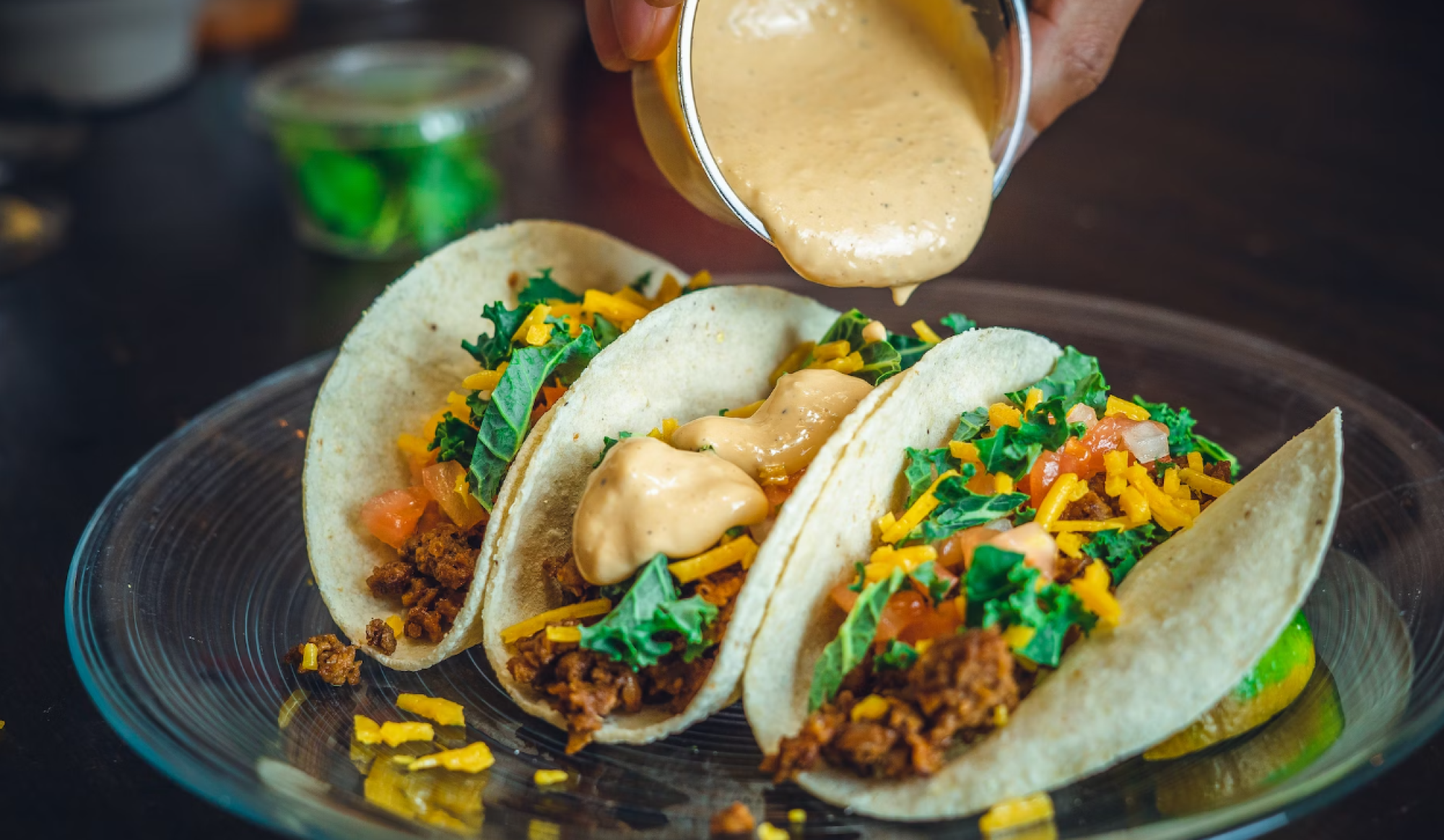 The image shows three tacos filled with ground meat, chopped vegetables, shredded cheese, and sauce being poured over them, served on a glass plate.