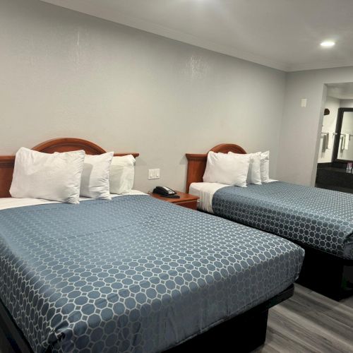 The image shows a tidy hotel room with two double beds, blue bedspreads, wooden headboards, and nightstand, plus a mirror and small counter.