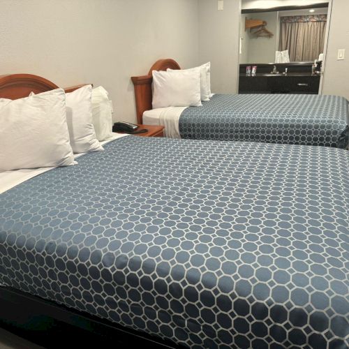 This image depicts a hotel room with two double beds covered in blue hexagonal patterned bedspreads, white pillows, and a nightstand between them.