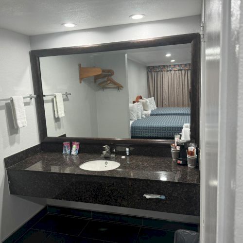 The image shows a bathroom vanity with a sink, towels, and toiletries. There's a large mirror reflecting a bedroom with a bed and hanging clothes.