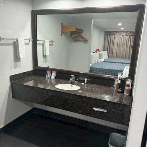 A bathroom vanity with a large mirror, a sink, toiletries, and towels. In the reflection, a bedroom with two beds and a window is visible.