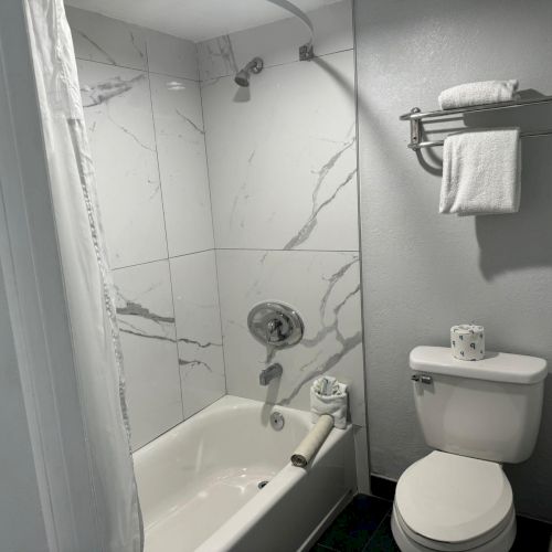 This image shows a modern bathroom with a marble-tiled bathtub, a shower, a toilet, and a towel rack holding towels above the toilet.