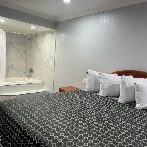 A hotel room with a bed featuring a black and white hexagonal patterned bedspread, several pillows, a nightstand, and an open bathroom door.