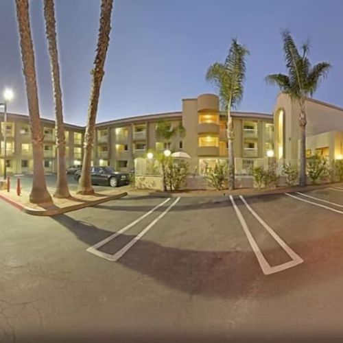 A three-story hotel building with palm trees in the parking lot during dusk, showcasing empty parking spaces and lit exterior corridors.