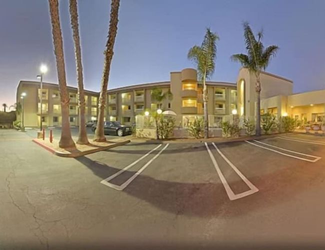 A three-story hotel building with palm trees in the parking lot during dusk, showcasing empty parking spaces and lit exterior corridors.