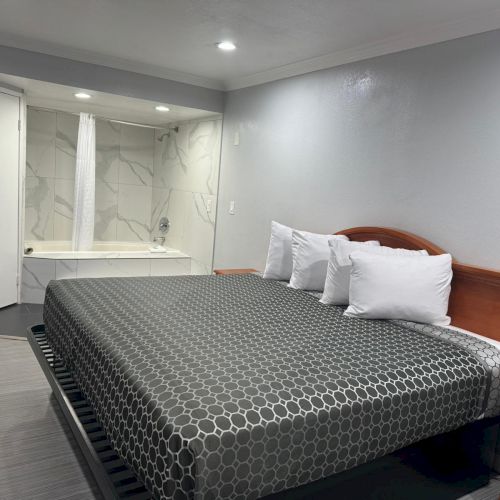 A modern hotel room with a large bed, white sheets, a wooden headboard, and a corner bathtub near a white door.