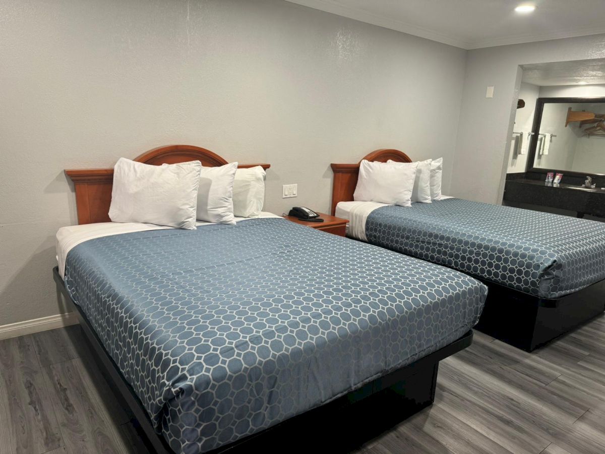 The image shows a hotel room with two neatly made beds, each with blue-patterned bedspreads and white pillows, on a wooden floor, and a nightstand in between.