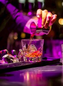 A bartender is pouring a drink into a glass with ice at a bar, with other professional bar tools visible and a second glass on the counter.