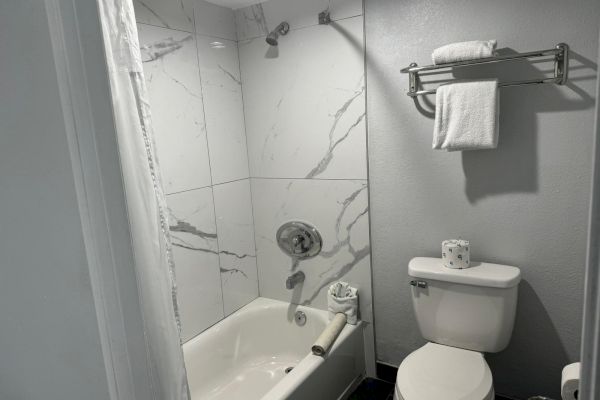 This image shows a bathroom with a bathtub, a showerhead, a toilet, and a towel rack holding folded towels above the toilet, always ending the sentence.