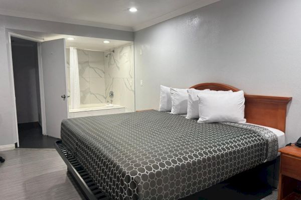 A modern hotel room with a king-size bed, three pillows, nightstand, and an open bathroom area with a bathtub and shower.