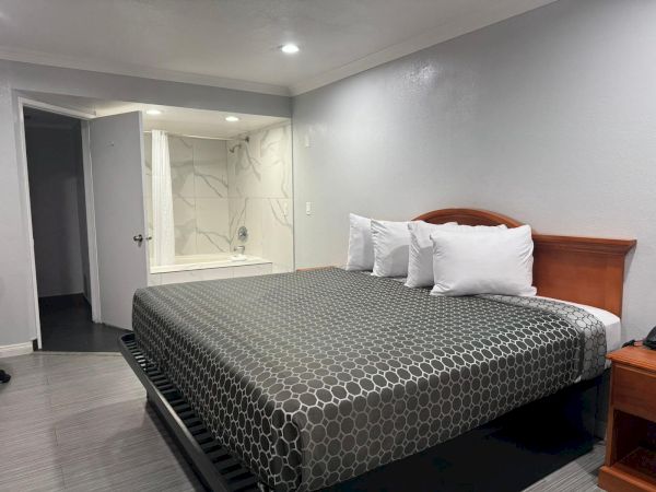 A modern hotel room with a king-size bed, three pillows, nightstand, and an open bathroom area with a bathtub and shower.
