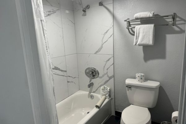 The image shows a small bathroom with a tiled bathtub, a toilet, and a towel rack holding folded towels and a roll of toilet paper.