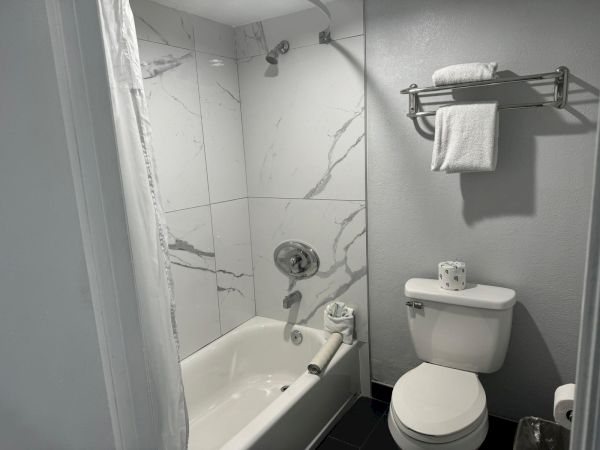 The image shows a small bathroom with a tiled bathtub, a toilet, and a towel rack holding folded towels and a roll of toilet paper.