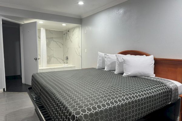 A tidy hotel room features a large bed with a geometric-patterned cover, several pillows, and an adjacent open bathroom with a bathtub.