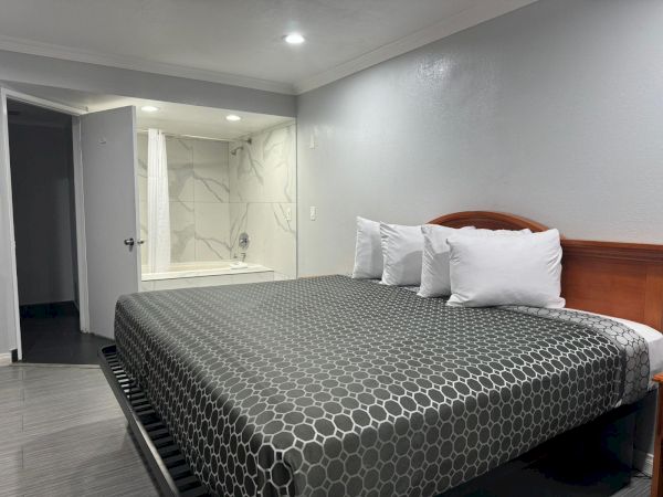 A tidy hotel room features a large bed with a geometric-patterned cover, several pillows, and an adjacent open bathroom with a bathtub.