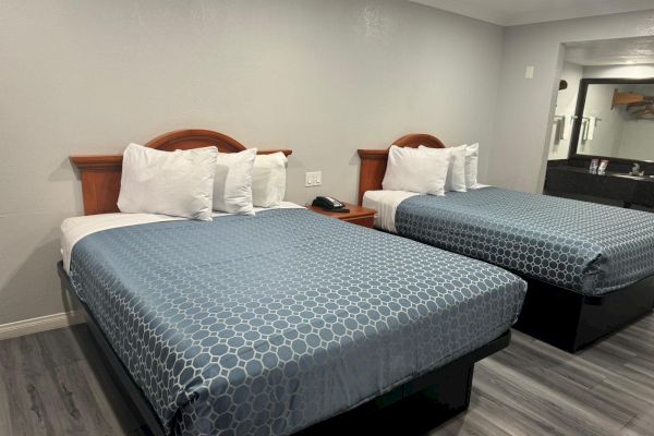 The image shows a hotel room with two neatly made beds, blue patterned bedspreads, and white pillows. There's a bedside table with a phone.